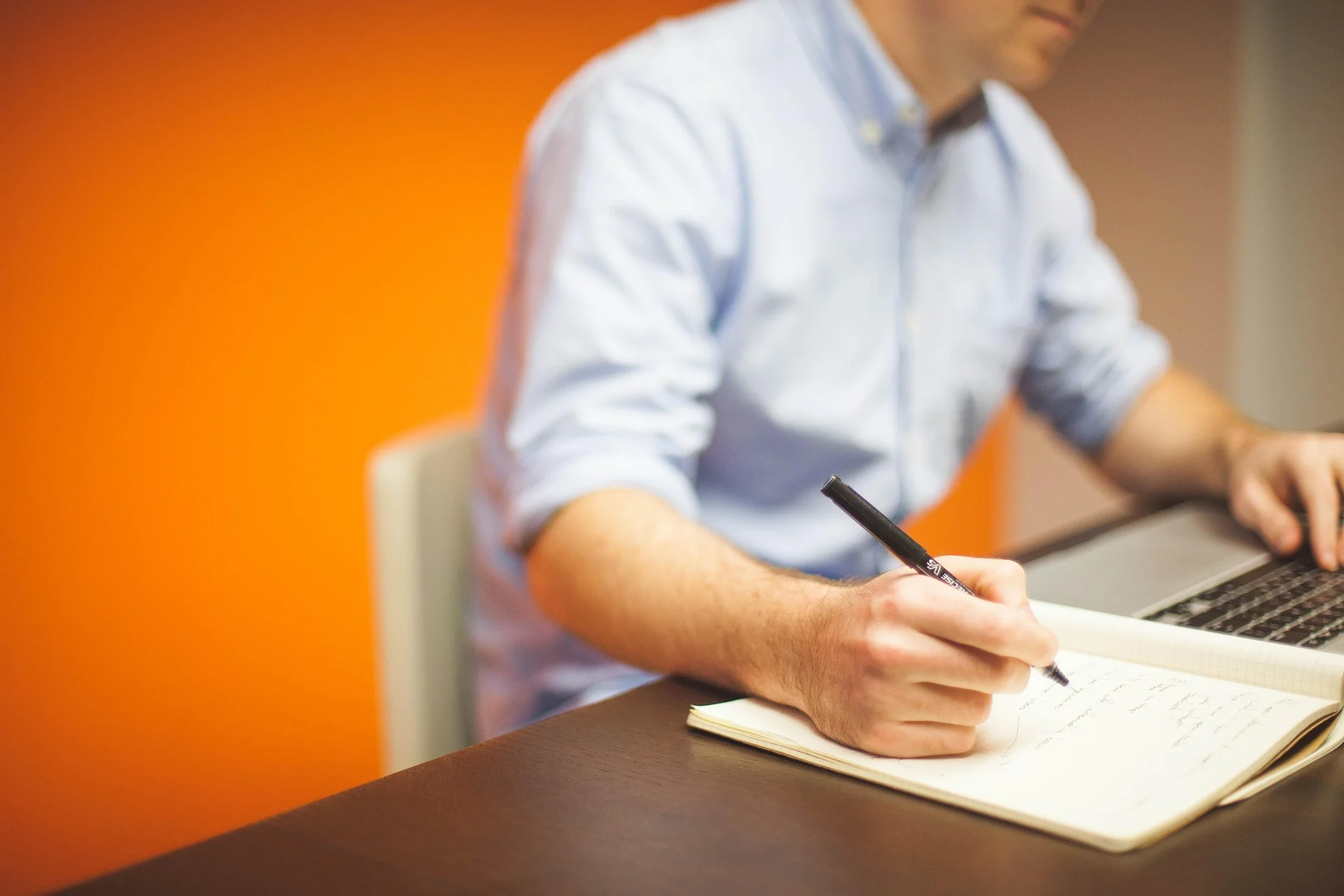Person writing notes at laptop workspace.
